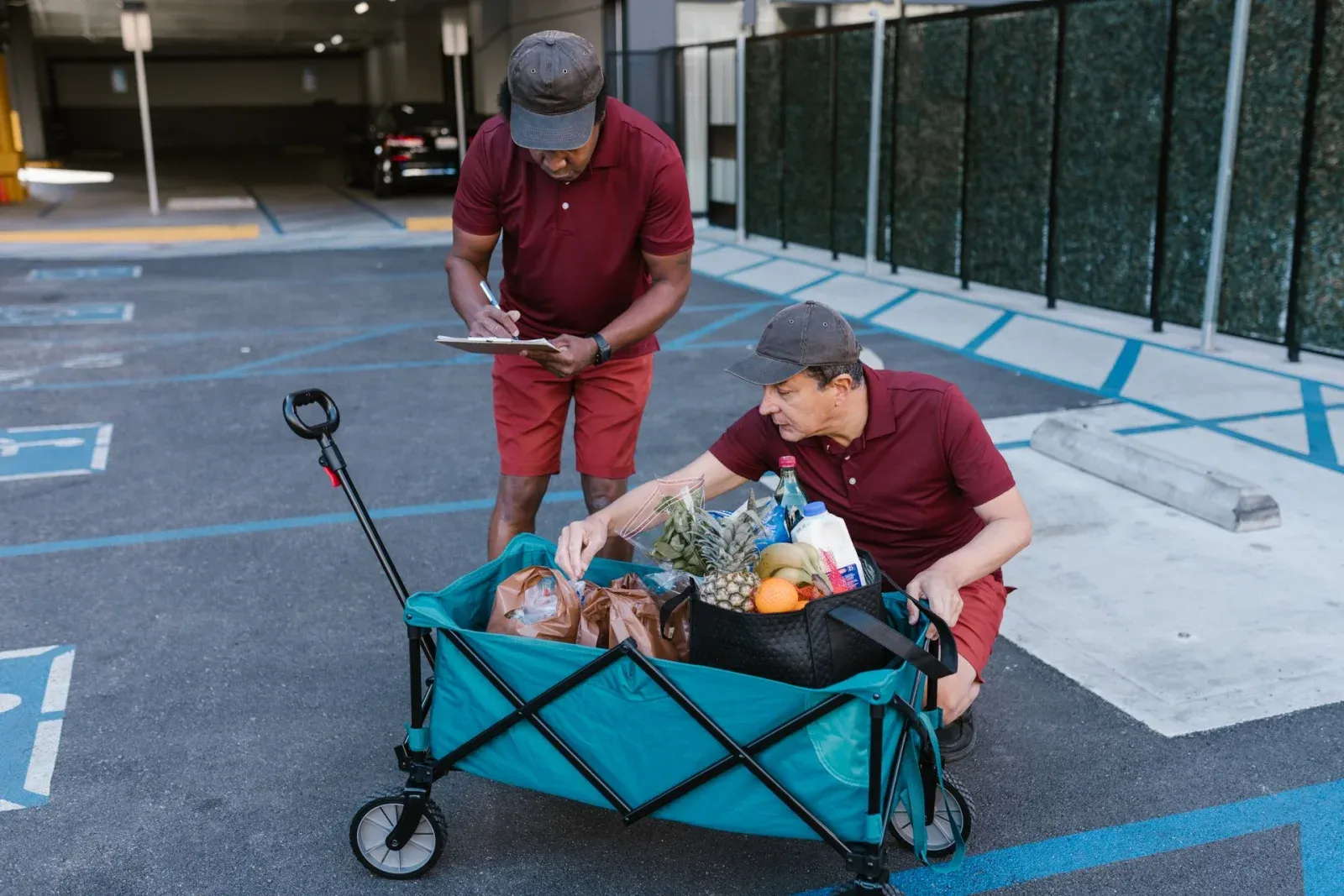 Two workers checking groceries beside a cart, with one recording notes on a clipboard.